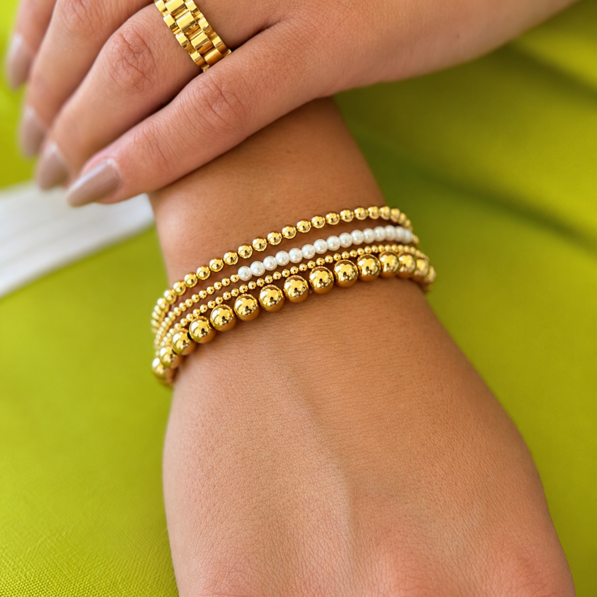 A close-up of a person's wrist wearing a stack of gold beaded bracelets, including textured round beads and smooth pearl-like beads, with a matching gold ring on the finger. The background is a bright lime green fabric.