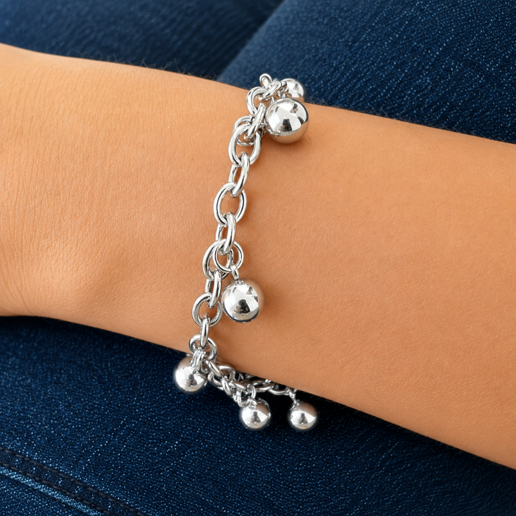 A silver charm bracelet with small ball charms and a dangling chain, shown worn on a person's wrist against a dark blue denim background.