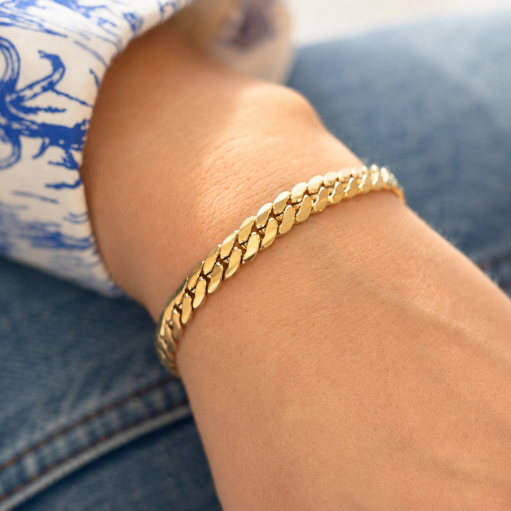 A close-up of a person's wrist wearing a shiny gold curb link bracelet, resting against blue denim jeans and a white shirt with a blue graphic print.