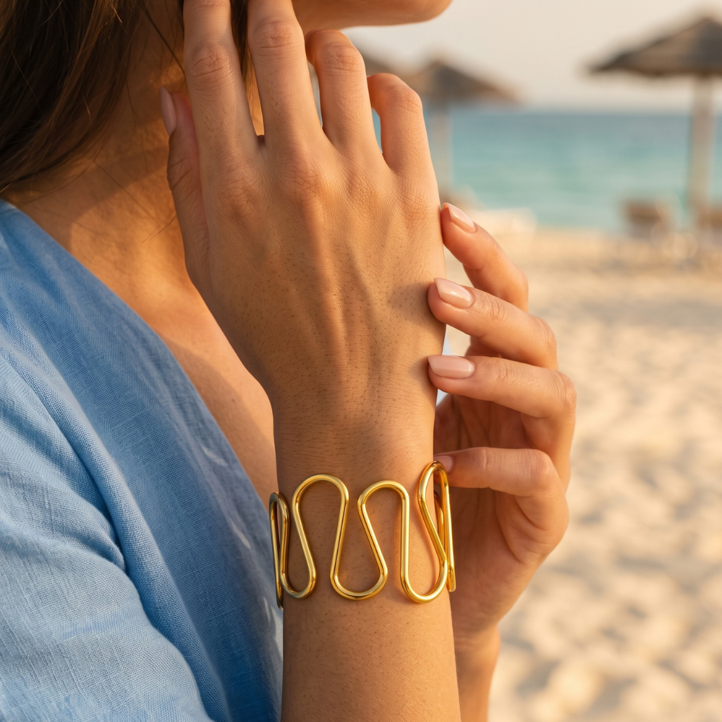 A gold wire cuff bracelet worn on a woman's wrist, shown in a close-up beach lifestyle shot with sand and ocean in the background.