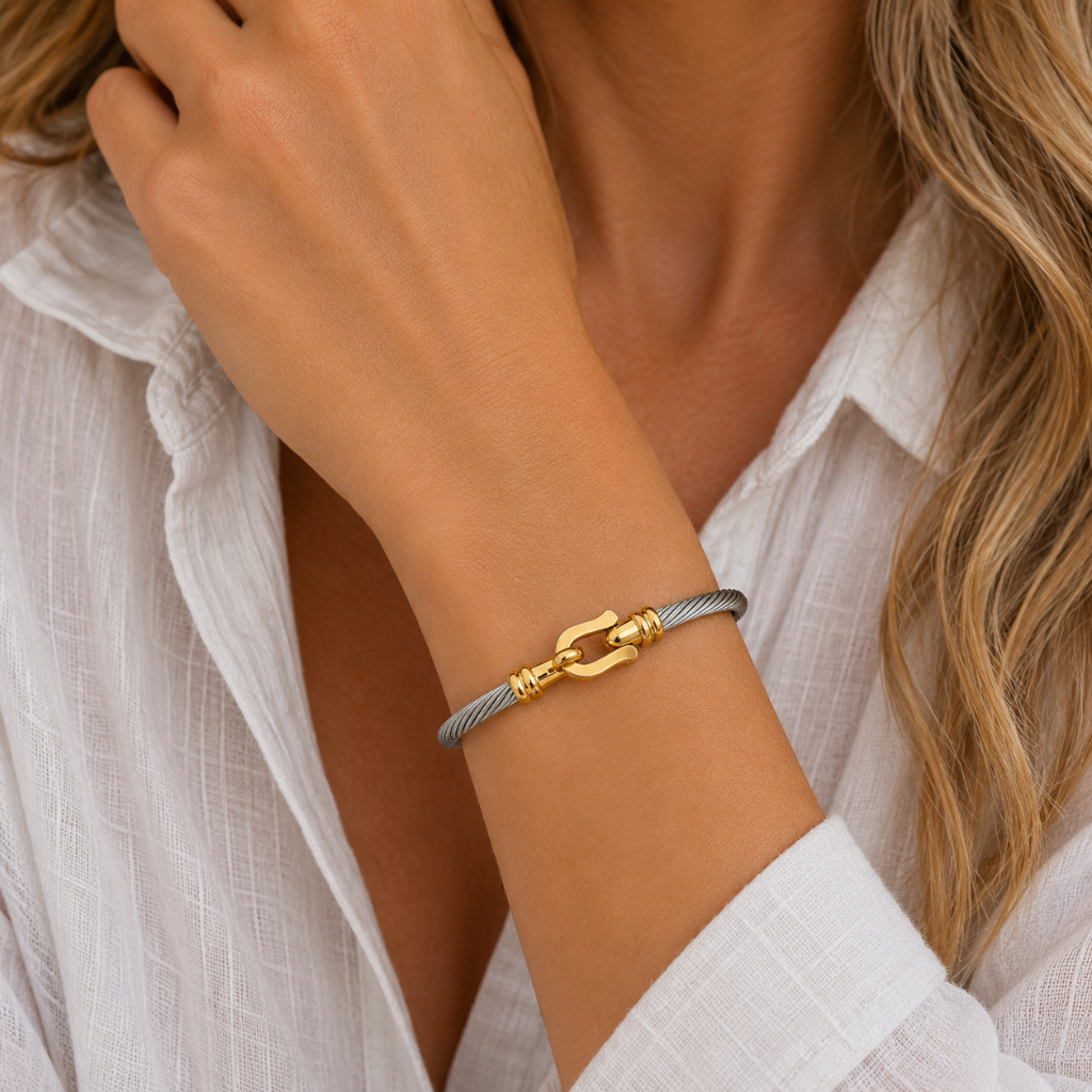 A close-up of a woman's wrist wearing a delicate gray braided cord bracelet with a polished gold clasp, paired with a white button-down shirt.