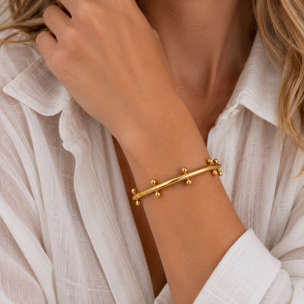 A close-up of a woman wearing a delicate gold bangle bracelet with small bead accents, paired with a white textured blouse.