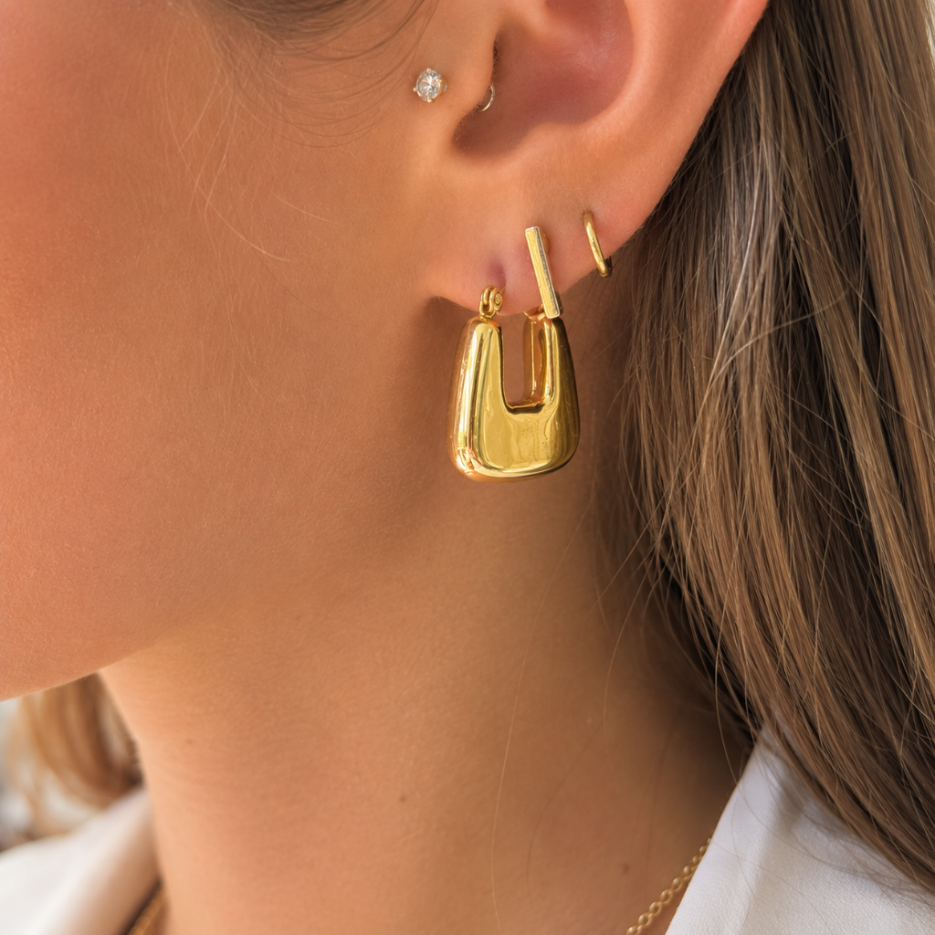 A close-up of a woman's ear wearing a polished gold hoop earring with a curved, open U-shaped design. The image highlights the gold jewelry against her skin and hair.