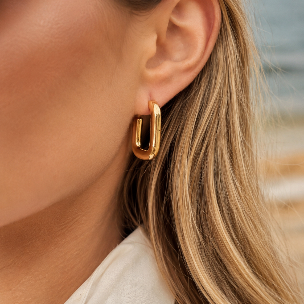 A close-up of a woman's ear wearing a polished gold geometric hoop earring, with her blonde hair and a white top visible against a softly blurred outdoor background.