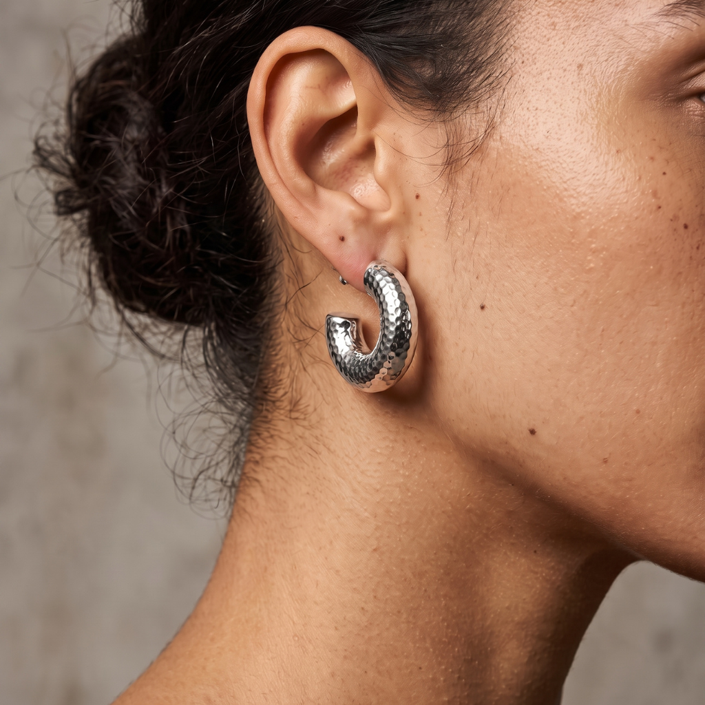 A close-up of a model's ear wearing a textured silver crescent-shaped hoop earring against a neutral gray background.