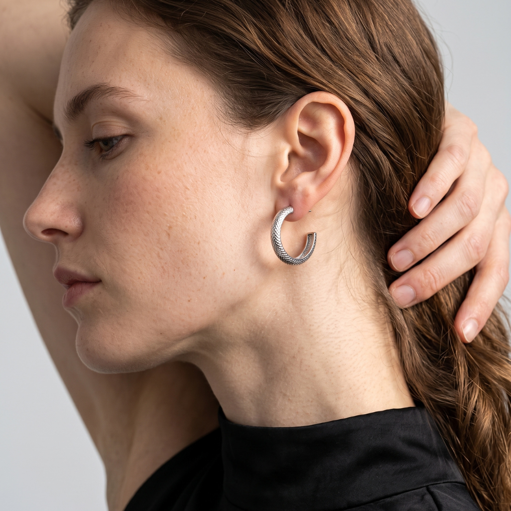 A close-up side profile of a woman wearing a textured silver C-shaped hoop earring, with her hand touching her hair against a neutral light gray background.