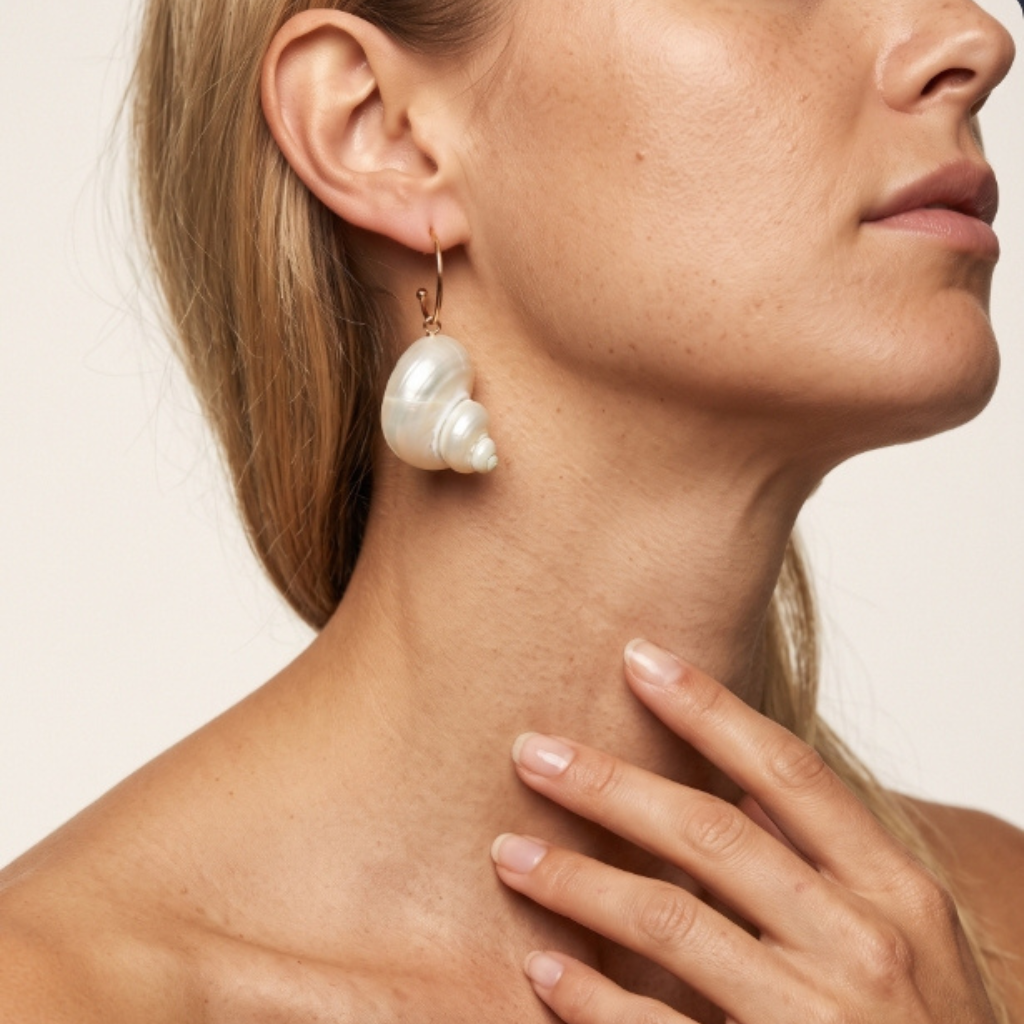 A close-up of a woman's neck and ear wearing a dangling shell-shaped pearl earring with a small gold hook.