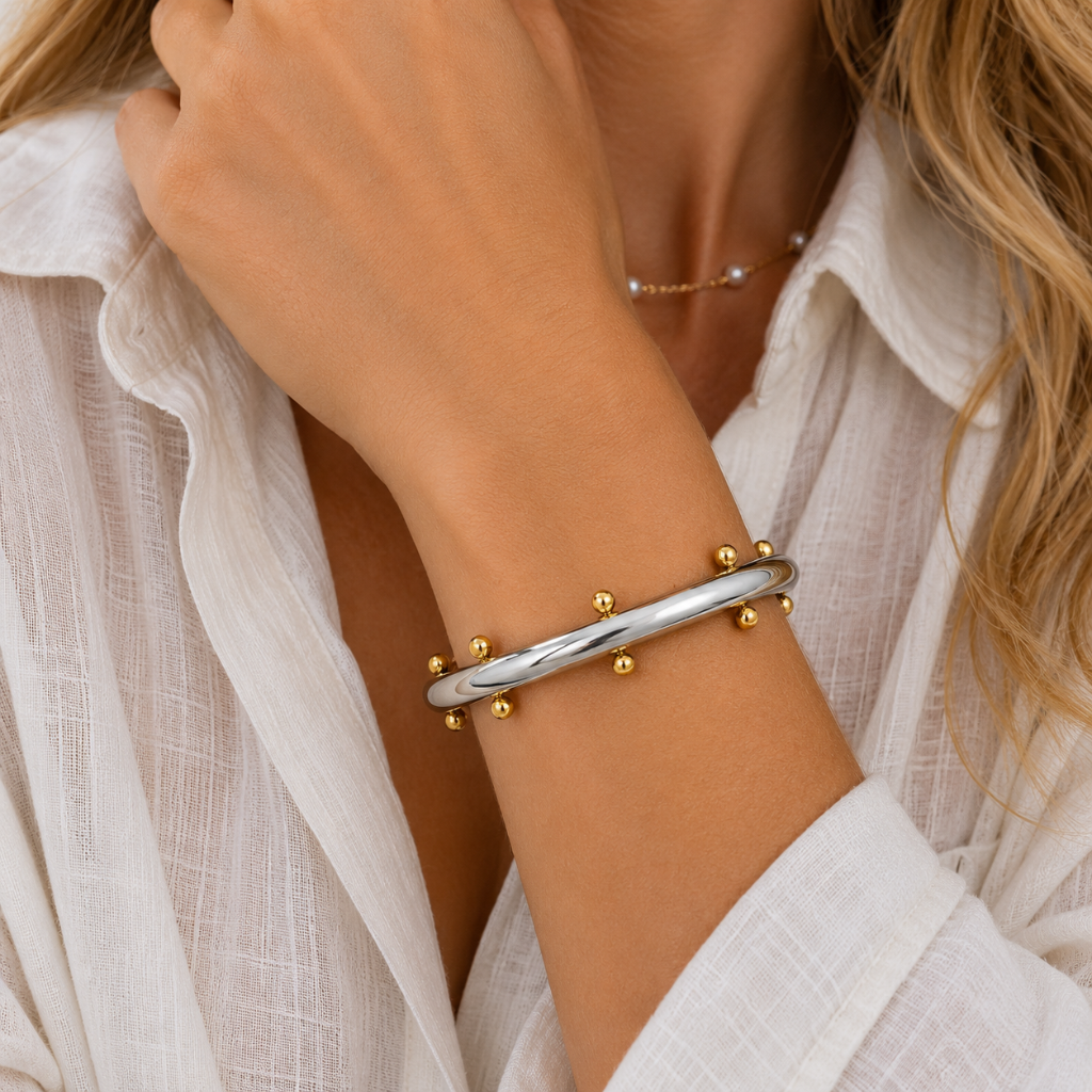 A close-up of a woman's wrist wearing a polished silver bangle bracelet with small gold ball accents, paired with a white textured blouse.
