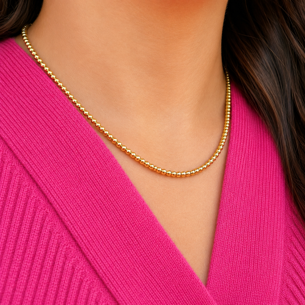 A close-up of a delicate gold beaded chain necklace worn by a model in a bright pink ribbed knit top.
