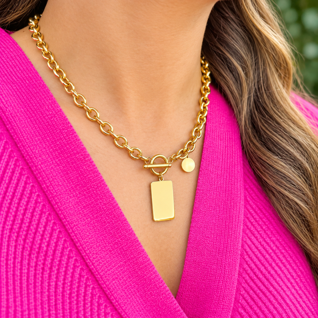 A gold chain necklace with a rectangular tag pendant and a small round charm, worn by a model in a bright pink ribbed top.