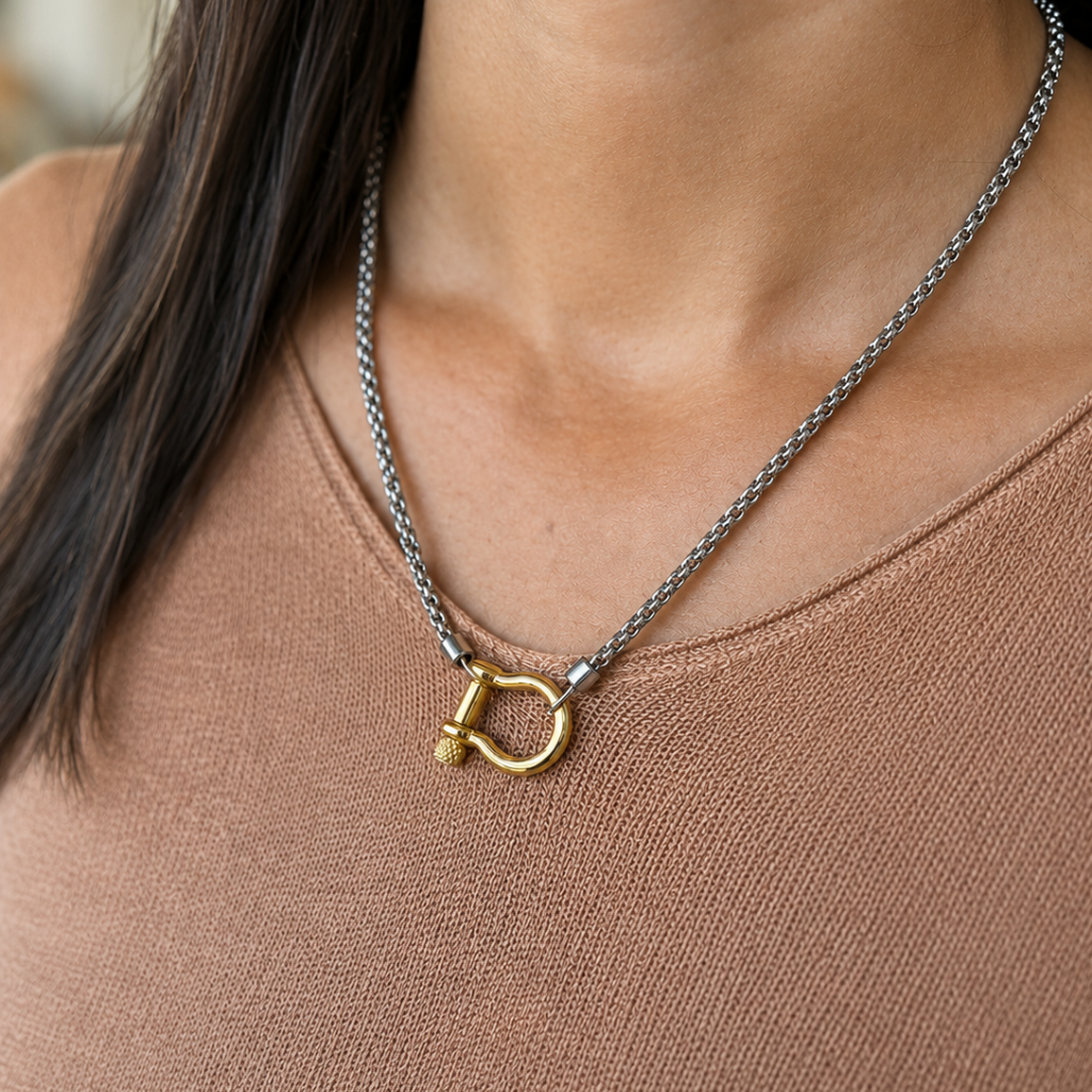 A close-up of a person wearing a silver-toned chain necklace with a gold-toned D-ring clasp and a small gold bead accent, paired with a brown knit top.