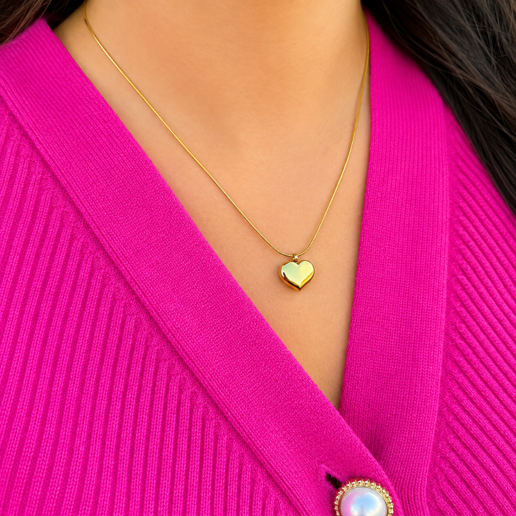 A close-up of a person wearing a delicate gold chain necklace with a small heart-shaped pendant, styled with a bright pink ribbed cardigan.
