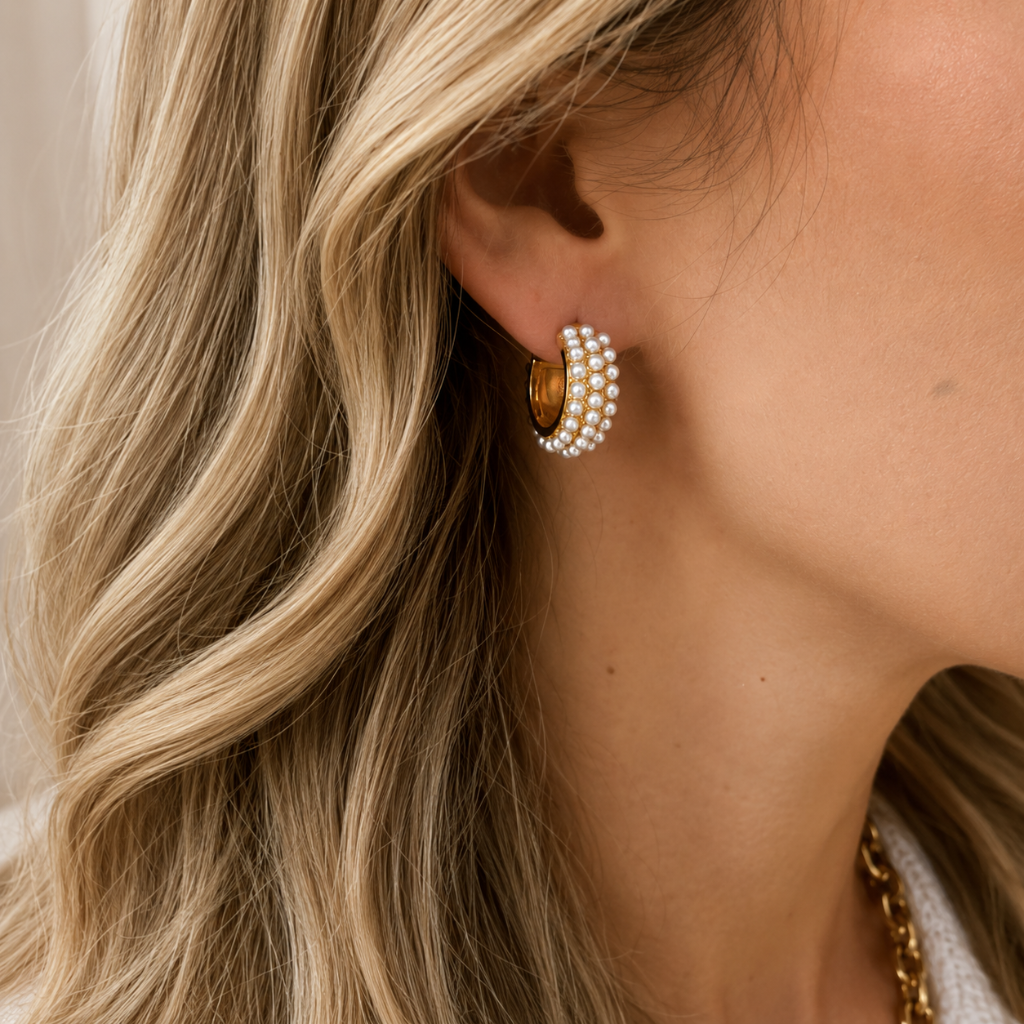 A close-up of a woman's ear wearing a gold hoop earring adorned with three rows of white pearls, set against her blonde hair and neck.