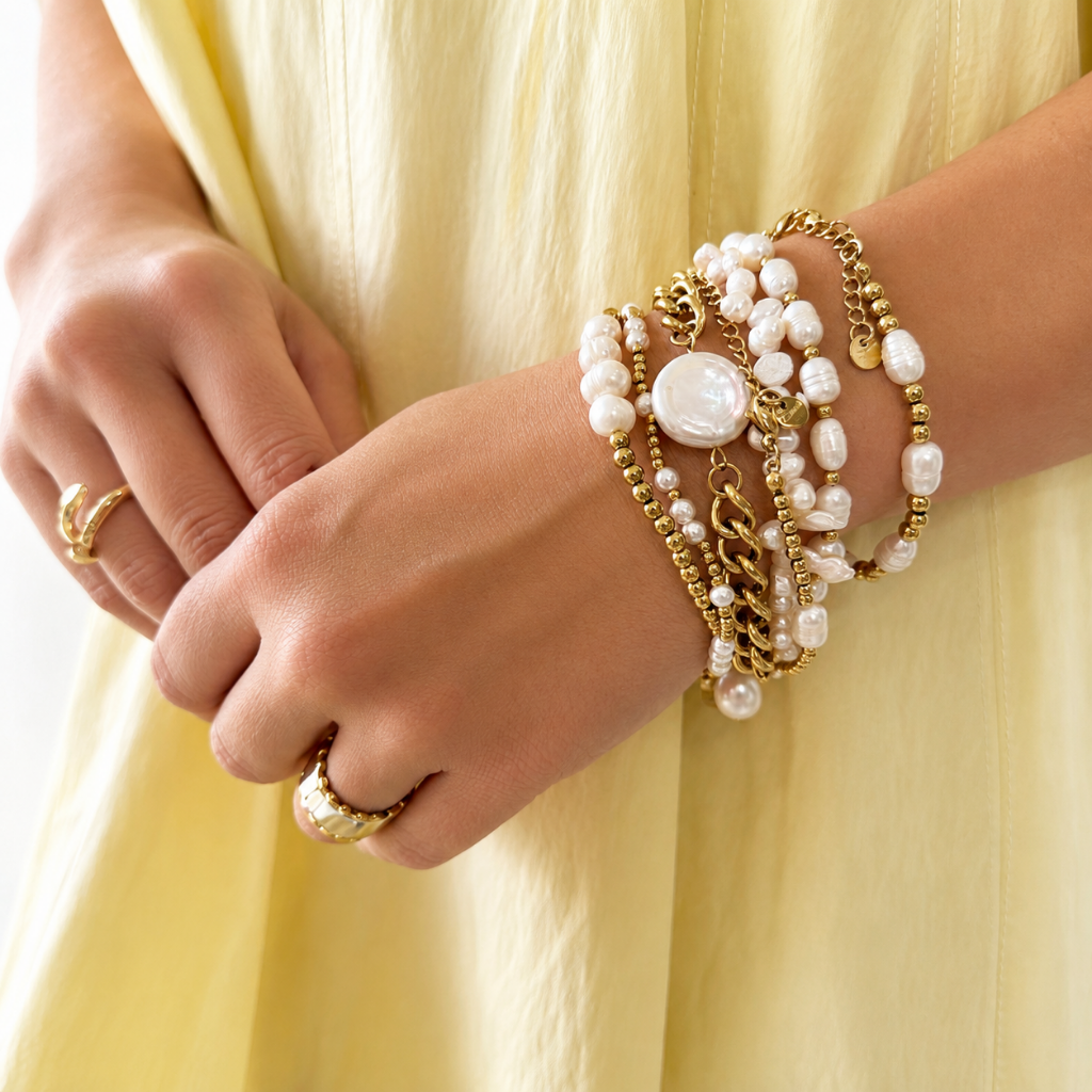 A close-up of a person's hands and wrist wearing a stack of gold and pearl bracelets, with matching gold rings, against a pale yellow dress.