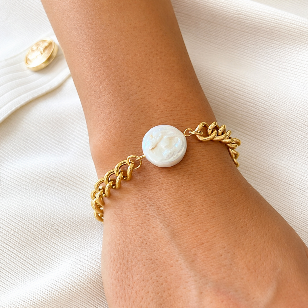 A gold chain bracelet with a round white cameo-style charm, worn on a person's wrist over a white textured top.