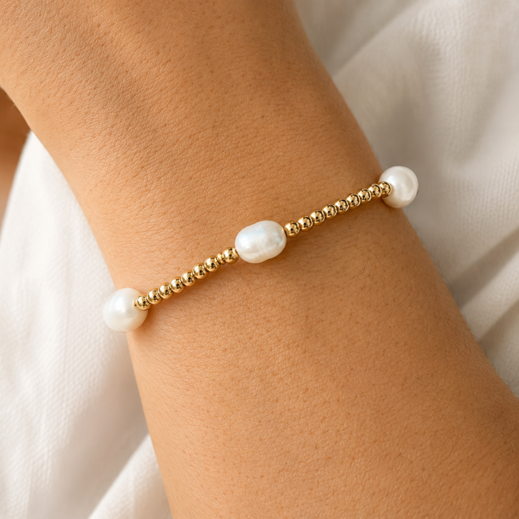A delicate gold beaded bracelet with three white pearl-like beads, shown worn on a person's wrist against a soft white fabric background.