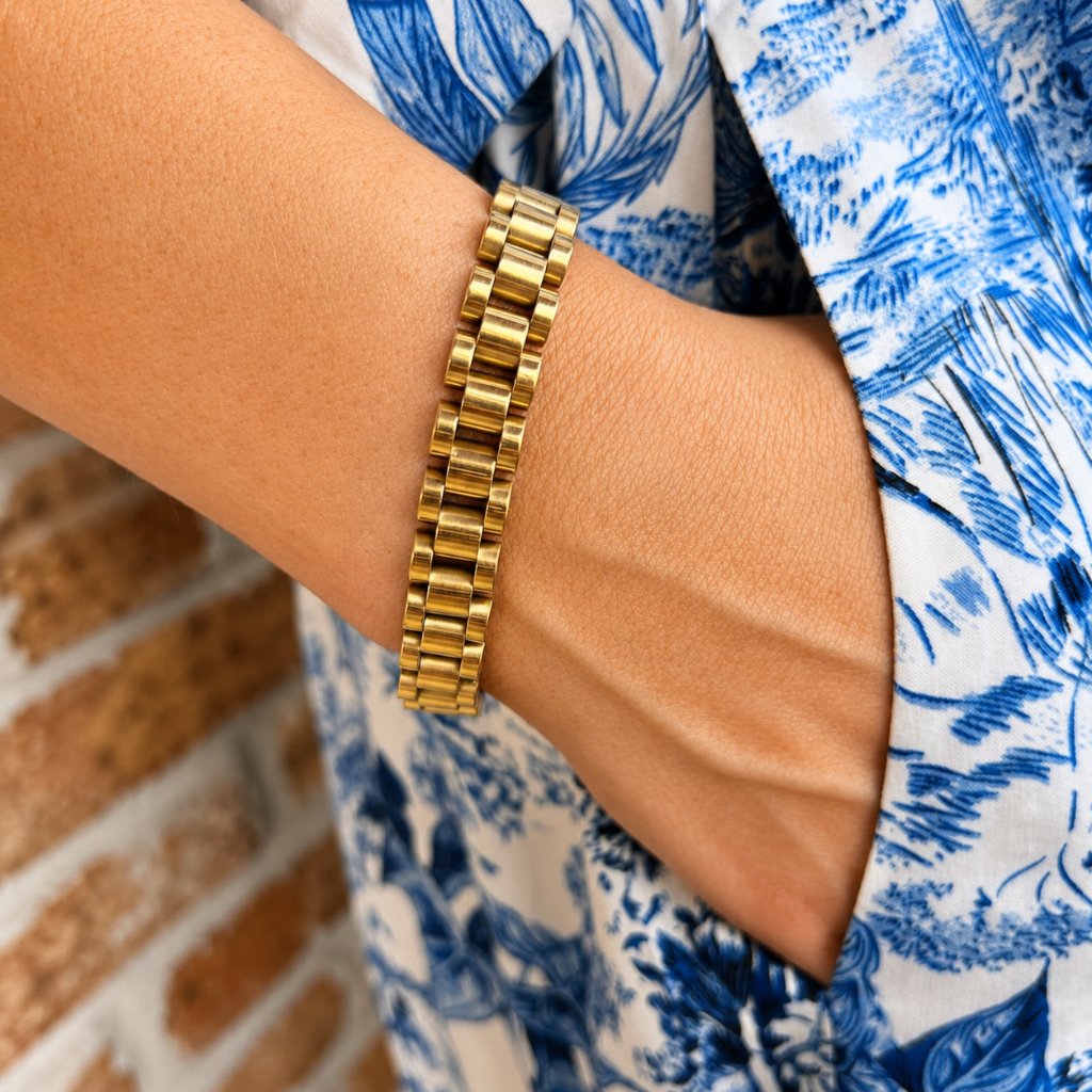 A close-up of a gold link bracelet worn on a person's wrist, with a blue and white patterned shirt and a brick wall in the background.