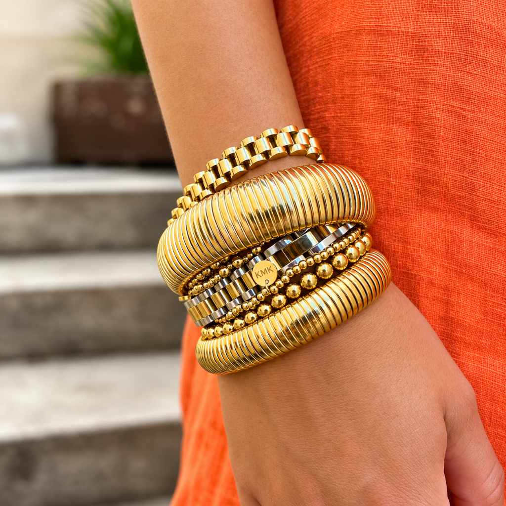 A close-up of a person's wrist wearing a stack of gold-toned bangles, including ribbed, beaded, and striped designs, paired with an orange sleeveless top.