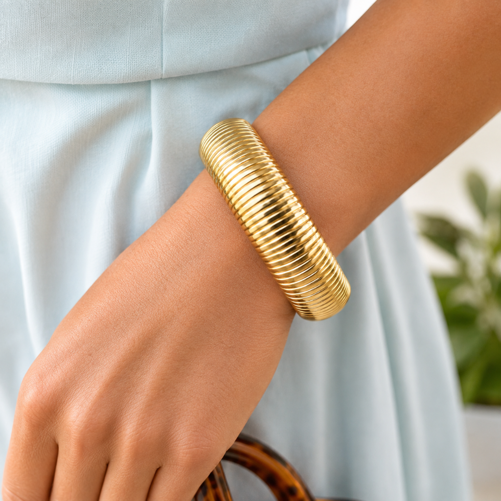 A close-up of a person's wrist wearing a wide, ribbed gold bangle bracelet, paired with a light blue dress.