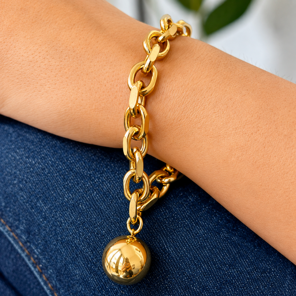 A close-up of a gold chain bracelet with a polished spherical charm, worn on a person's wrist over blue denim fabric.