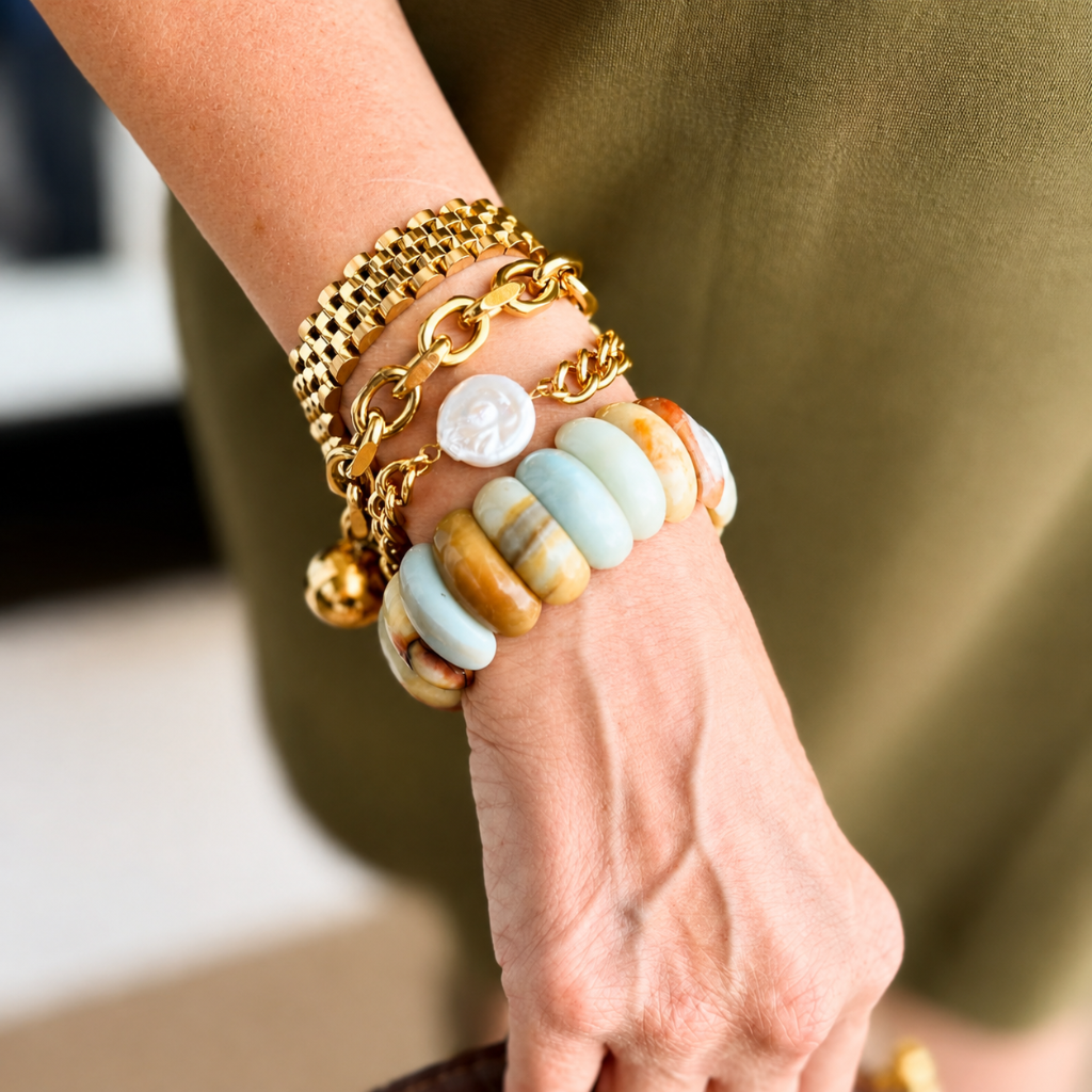 A close-up of a person's wrist and hand wearing multiple bracelets, including gold chain and mesh bracelets, a white shell charm, and a beaded bracelet with light blue, tan, and brown stones.
