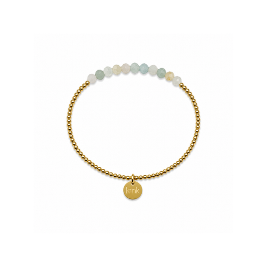 A delicate gold beaded bracelet with a small round gold charm engraved with 'kin', accented by a row of pale green and white beads at the top, shown on a plain white background.