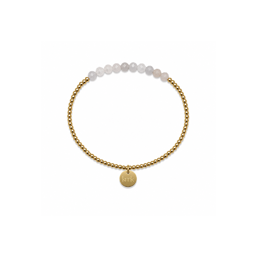 A delicate gold chain bracelet featuring a row of pale, translucent white beads at the top and a small round gold disc charm engraved with initials, set against a plain white background.
