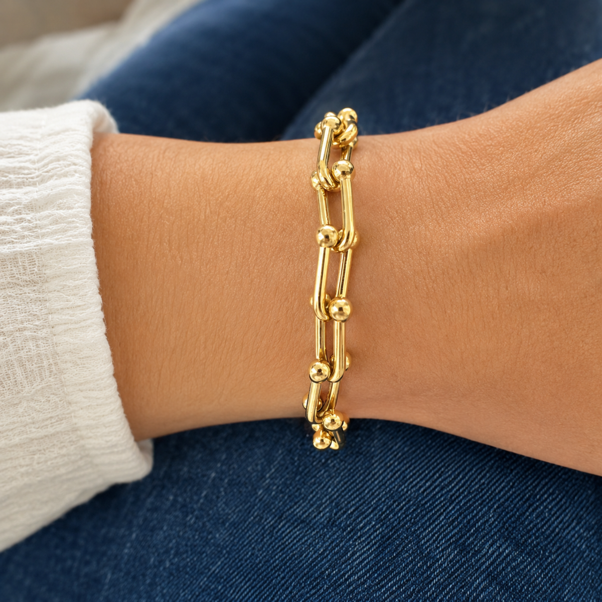 A close-up of a person's wrist wearing a gold chain bracelet with interlocking ball links, resting against blue denim jeans and a white sleeve.