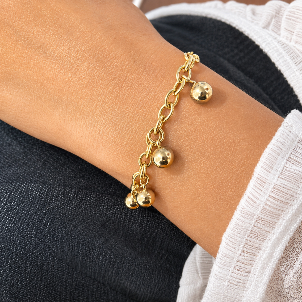 A close-up of a gold chain bracelet with three small polished ball charms worn on a person's wrist, shown against dark navy and white clothing.