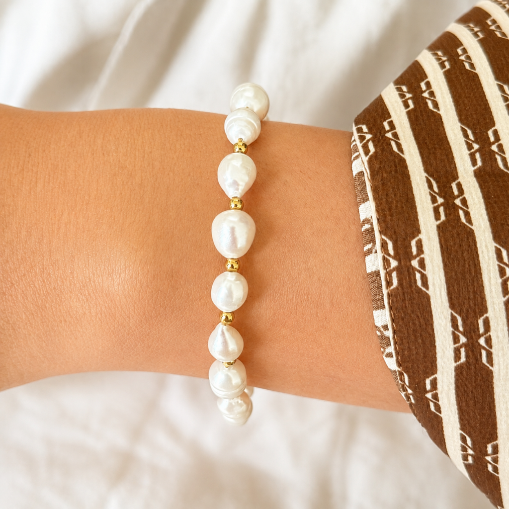 A close-up of a person's wrist wearing a beaded bracelet made of white baroque pearls separated by small gold spacer beads. The person is wearing a brown and cream patterned fabric sleeve, with a soft white draped fabric in the background.