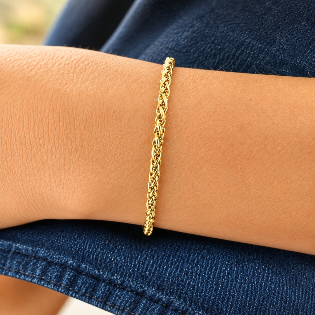 A close-up of a person's wrist wearing a thin braided gold chain bracelet, resting against dark blue denim fabric.