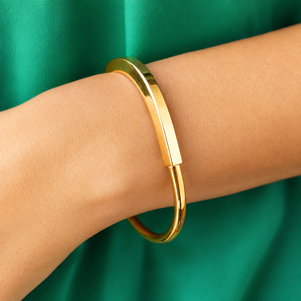 A polished gold bangle bracelet worn on a person's wrist, set against a vibrant green satin fabric background.
