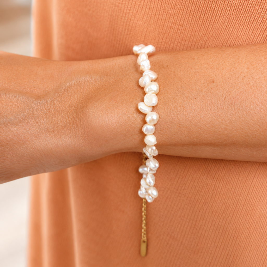A close-up of a person's wrist wearing a delicate bracelet made of irregular white baroque pearls and a small gold chain with a dangling tag, against a peach-colored knit top.