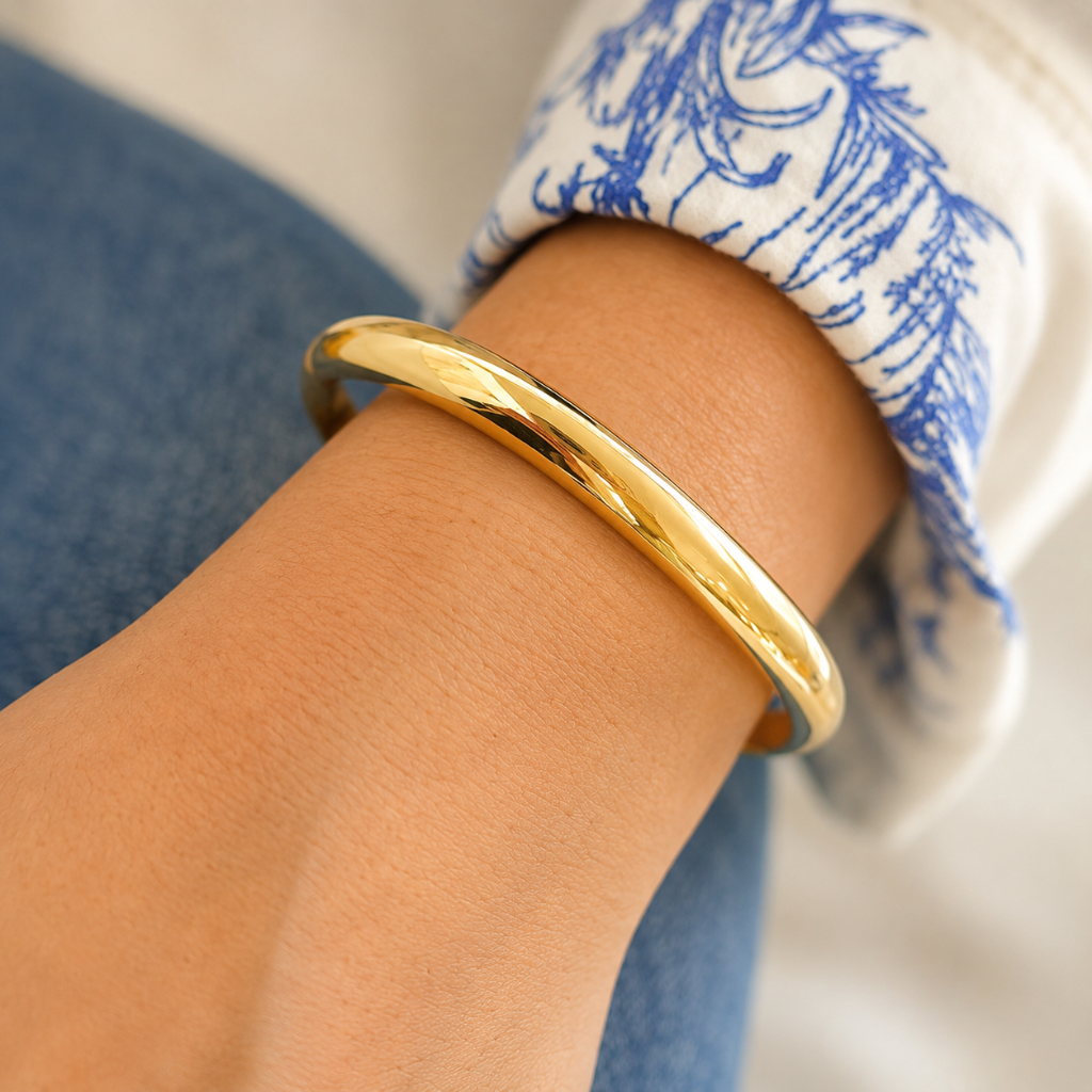 A polished gold bangle bracelet worn on a person's wrist, with a blue and white patterned cuff and blue jeans visible in the background.