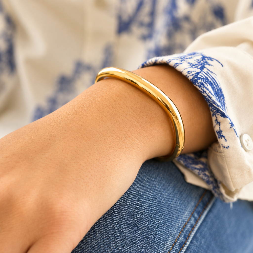 A polished gold bangle bracelet worn on a person's wrist, shown in a close-up lifestyle shot with a blue and white patterned shirt and blue jeans in the background.