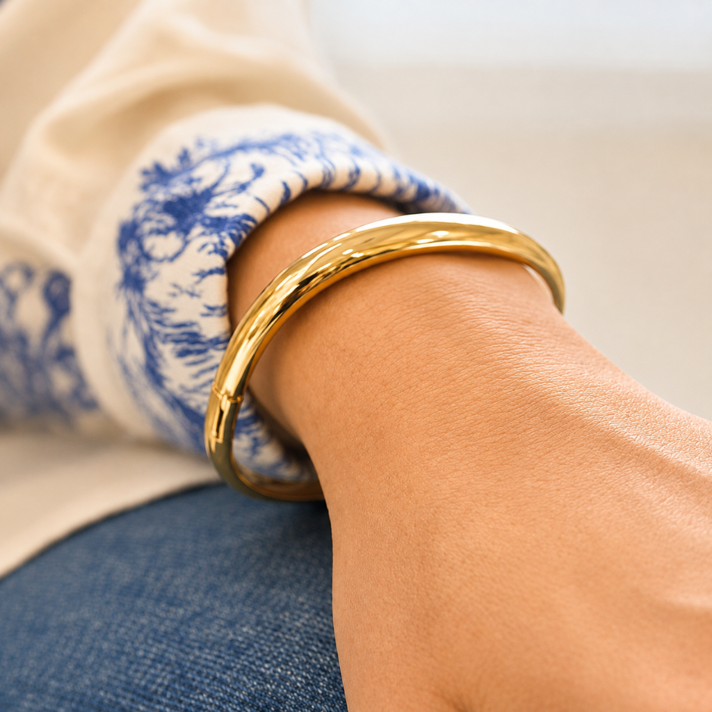 A polished gold bangle bracelet worn on a person's wrist, shown in a close-up lifestyle shot with a cream and blue patterned cuff and blue jeans visible.