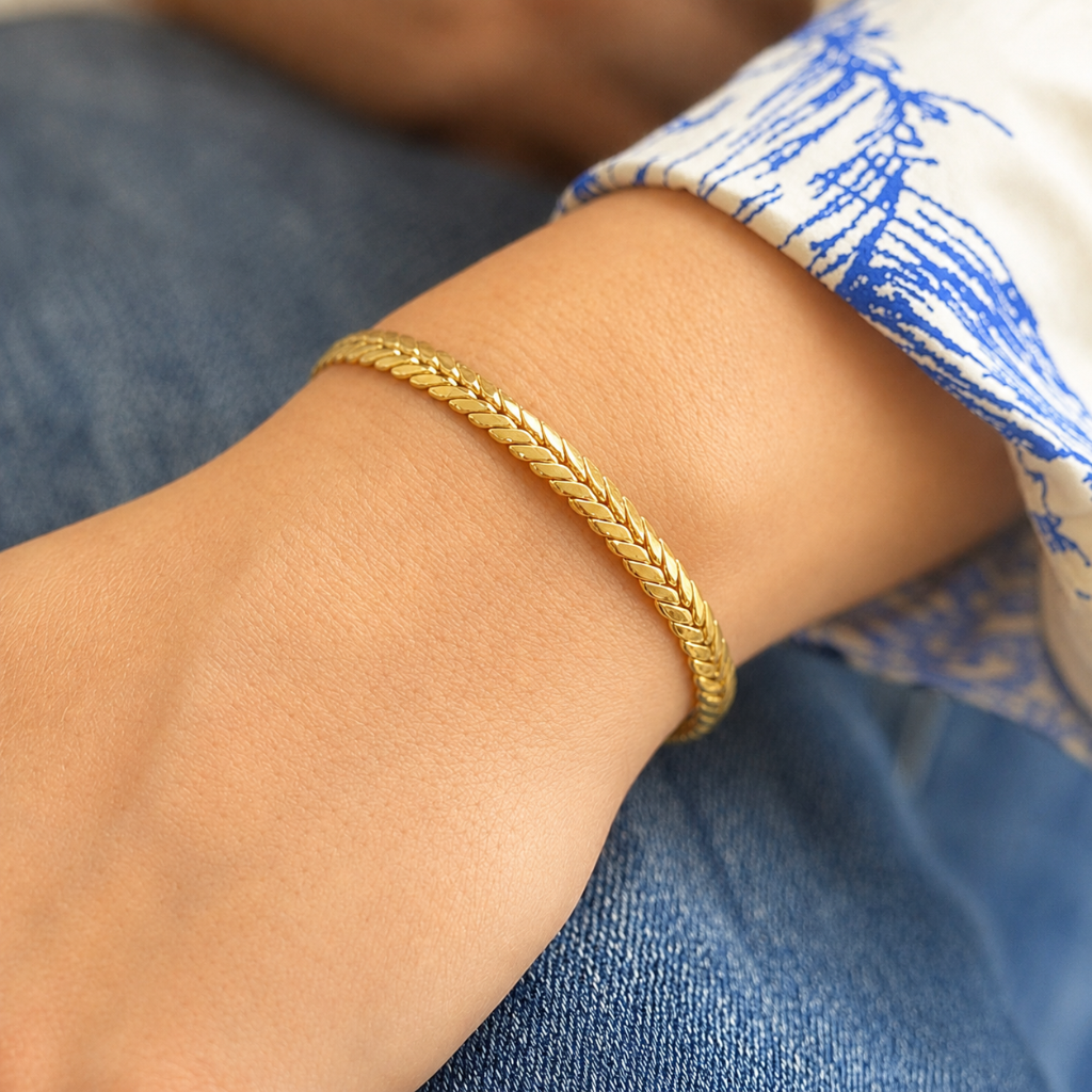 A close-up of a person's wrist wearing a thin braided gold chain bracelet, with blue jeans and a blue-and-white patterned shirt cuff visible.