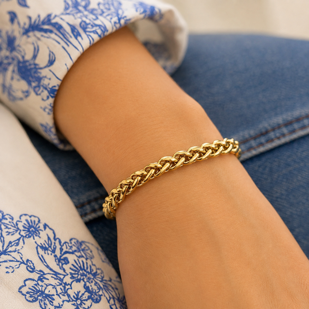 A close-up of a person's wrist wearing a polished gold curb chain bracelet, resting on blue denim jeans and a white blouse with a blue floral print.