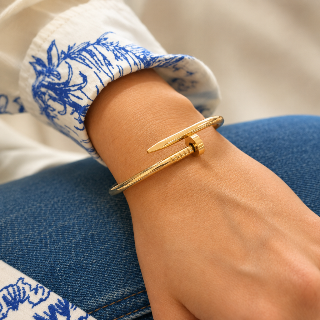 A gold-toned twisted bangle bracelet worn on a person's wrist, with a white and blue patterned sleeve and blue denim visible in the background.