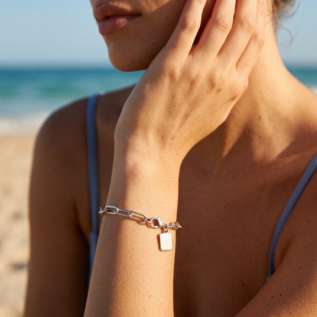 A silver chain bracelet with a small rectangular tag charm worn on a woman's wrist, photographed outdoors on a beach with the ocean and sand in the background.