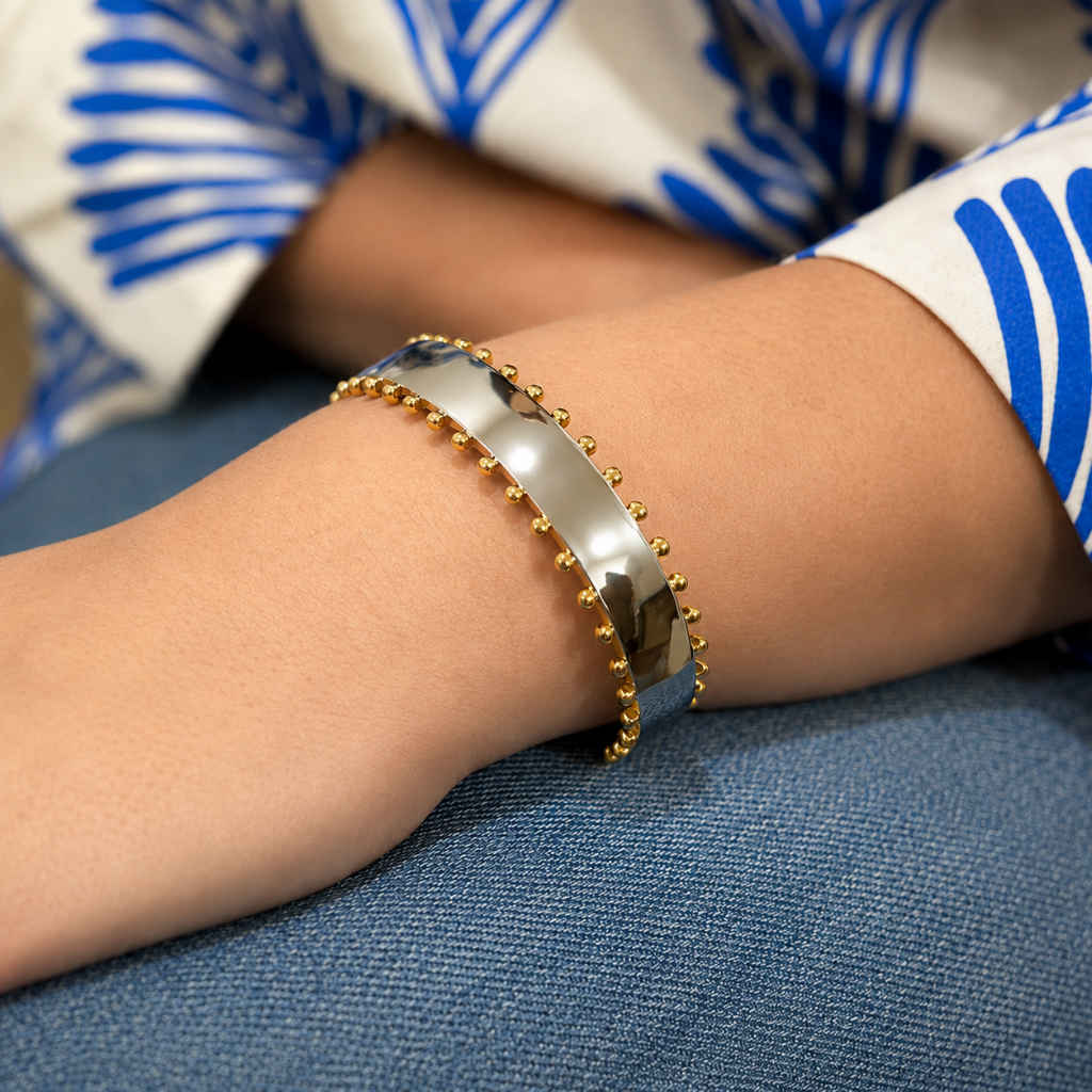 A close-up of a person's wrist wearing a polished silver bangle bracelet with small gold bead accents on the outer edge, resting on blue denim fabric.