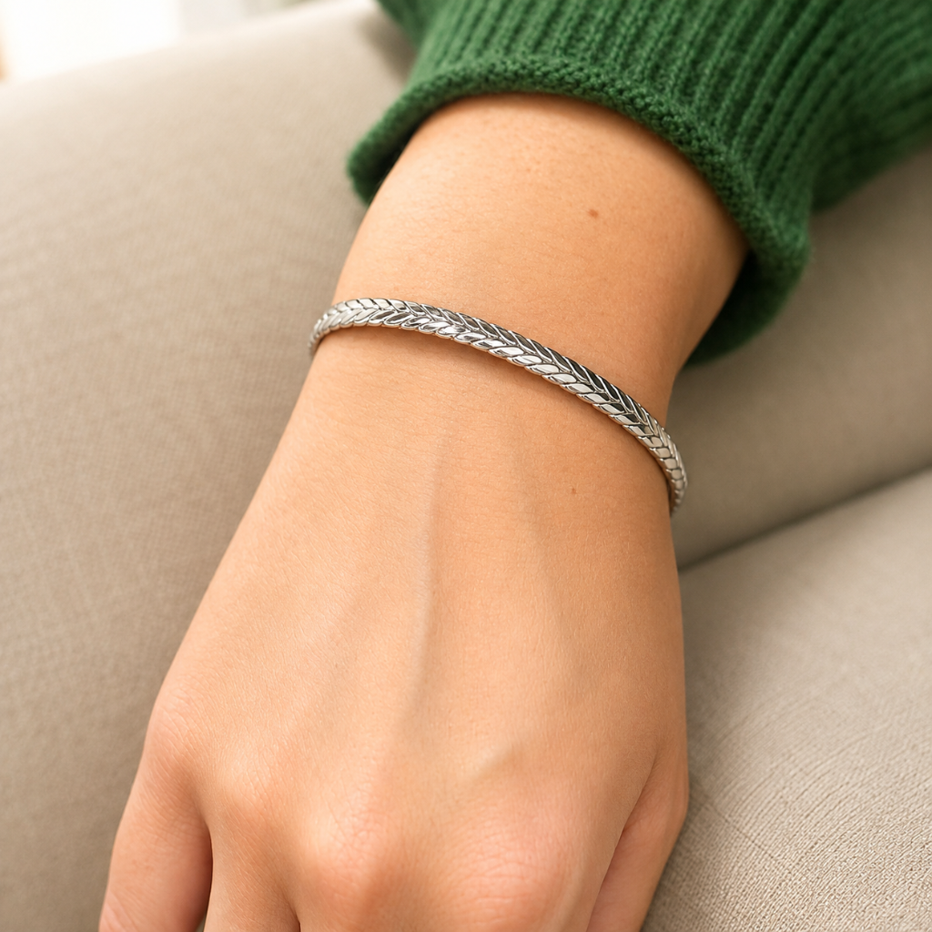 A close-up of a person's wrist wearing a textured silver braided bracelet, with a green knit sweater cuff visible and a beige upholstered chair in the background.