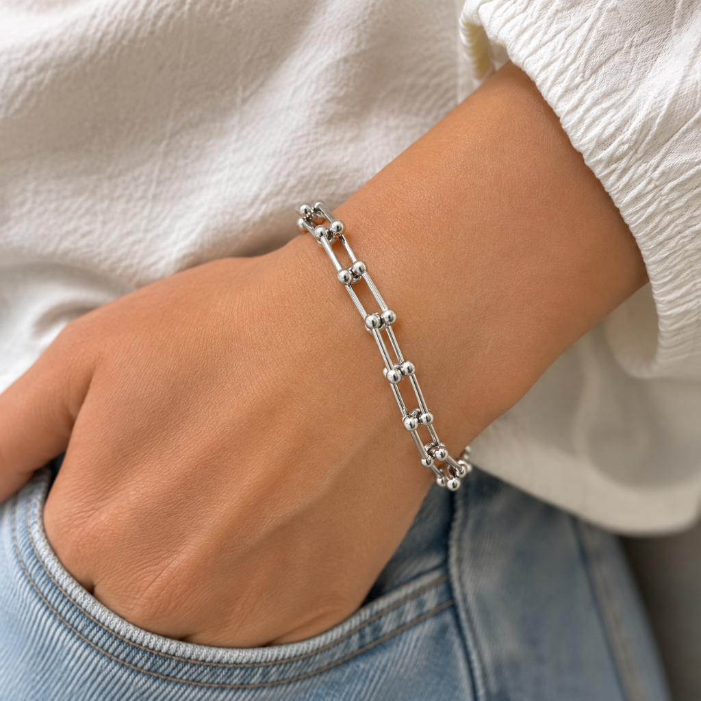 A close-up of a person's wrist wearing a silver chain bracelet with repeating double-bar links and small bead details, styled with a white textured top and blue jeans.