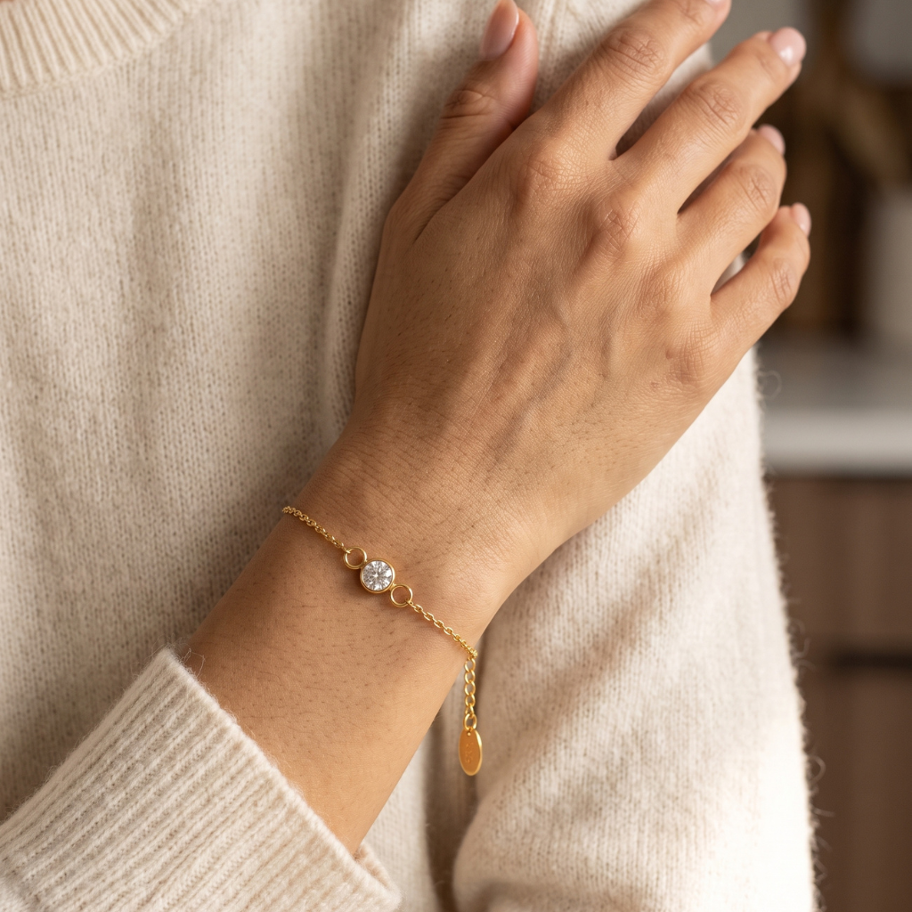 A delicate gold chain bracelet with a round clear gemstone and a small oval tag charm, shown worn on a person's wrist against a cream knit sweater.