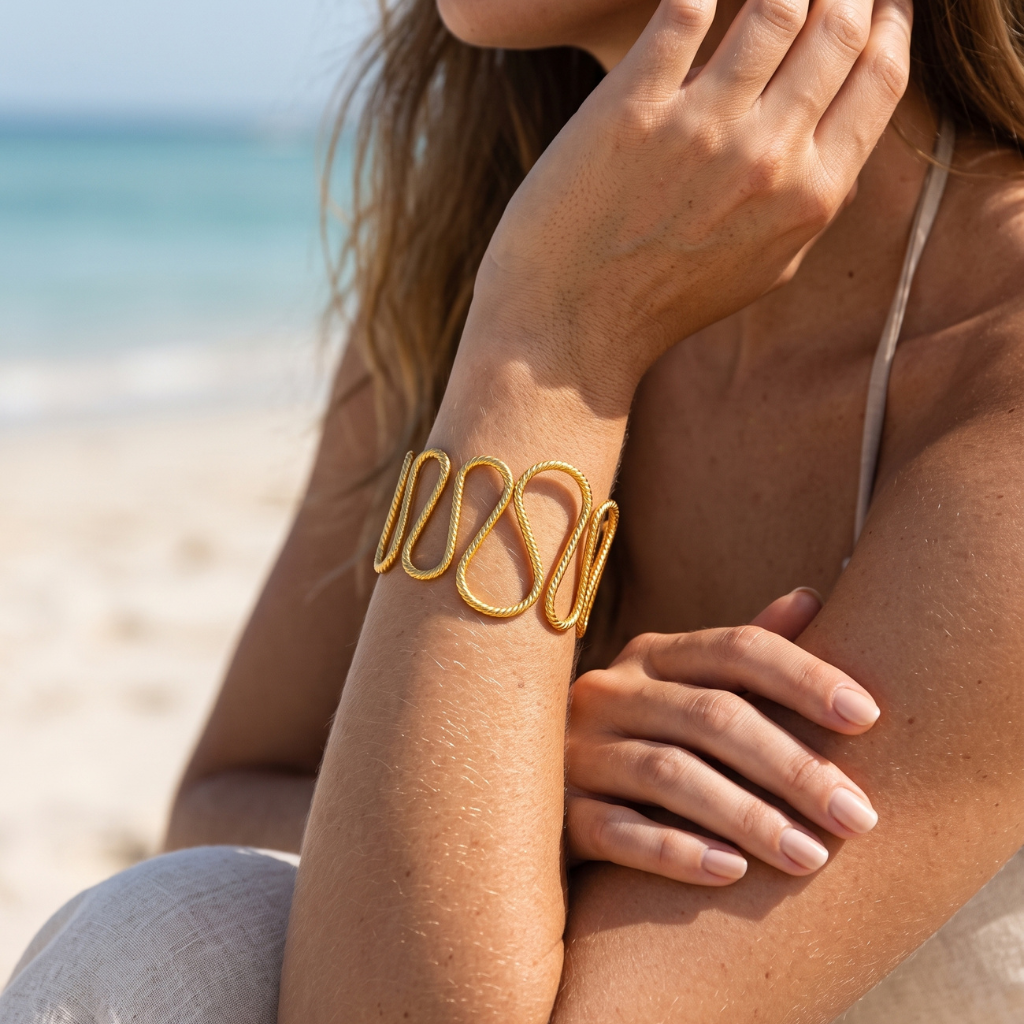 A gold-toned textured cuff bracelet worn on a woman's wrist, photographed on a beach with the ocean and sand blurred in the background.