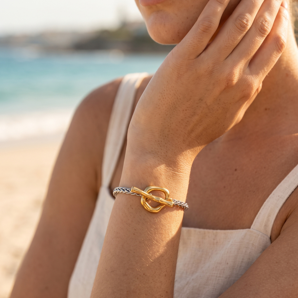 A close-up of a woman on a beach wearing a delicate bracelet with a gold interlocking rings design and a silver braided rope accent.