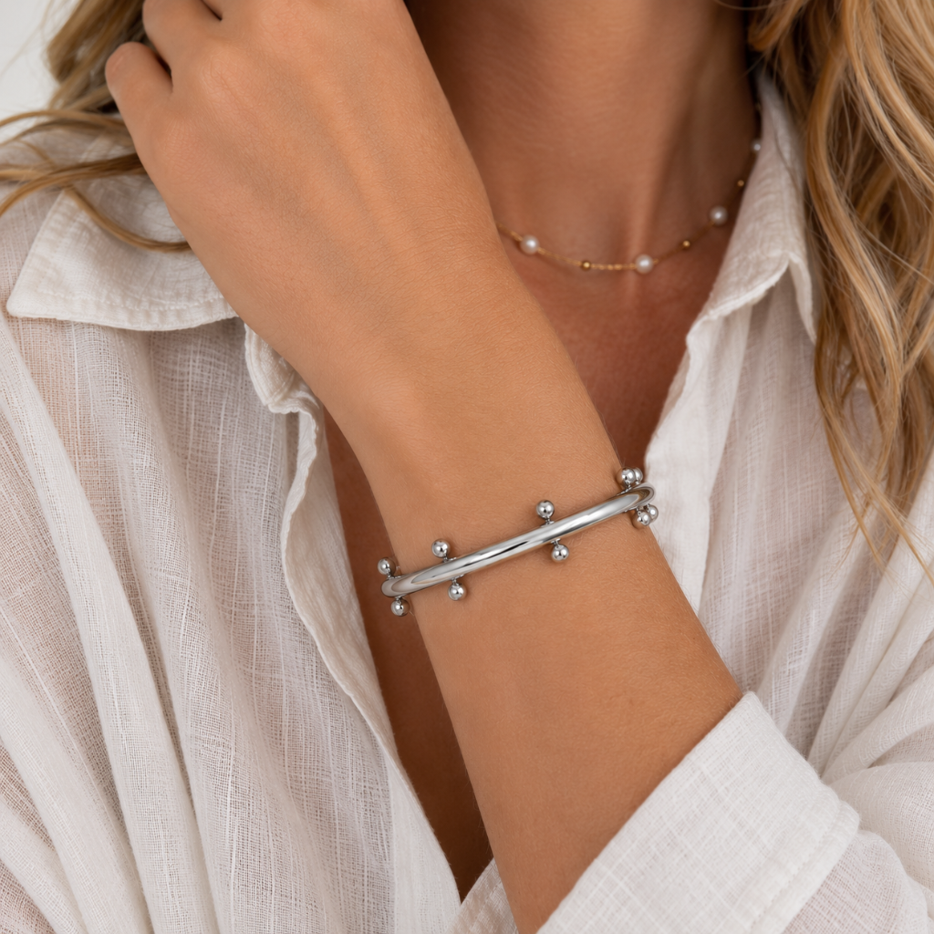 A close-up of a woman wearing a silver-toned bangle bracelet with small bead details, along with a delicate pearl necklace and a white button-up shirt.