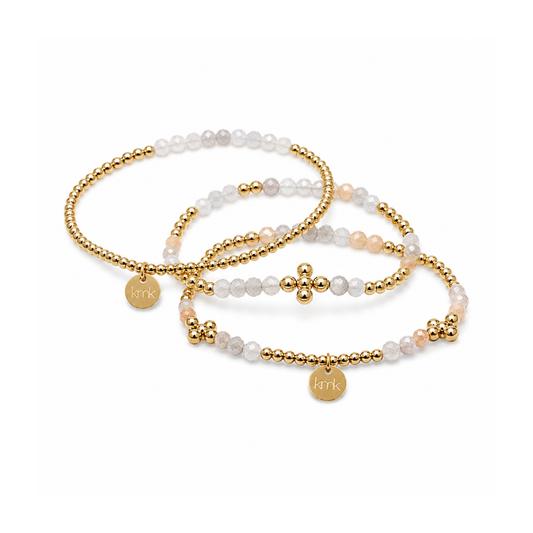 Three beaded stretch bracelets with gold-tone beads and charms, featuring white and pale peach beads, displayed on a clean white background.