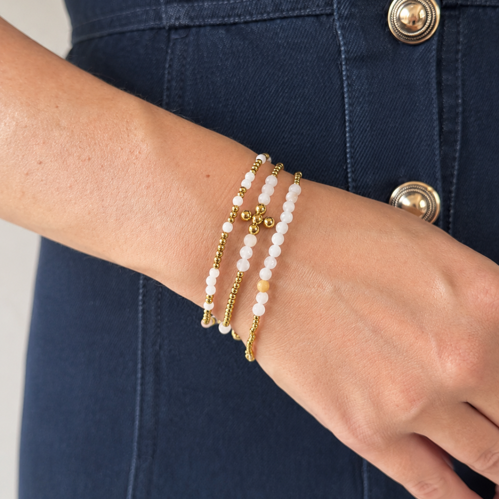 A close-up of a person's wrist wearing three delicate beaded bracelets: two strands of white pearl-like beads with gold spacer beads, and one gold beaded bracelet, shown against a dark blue denim top.