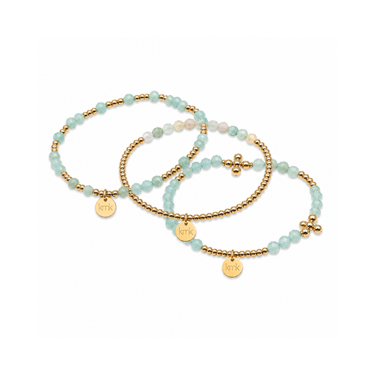Three beaded stretch bracelets with light blue-green stones, gold spacer beads, and gold-tone charms, including a cross charm and a round engraved disc charm, displayed on a clean white background.